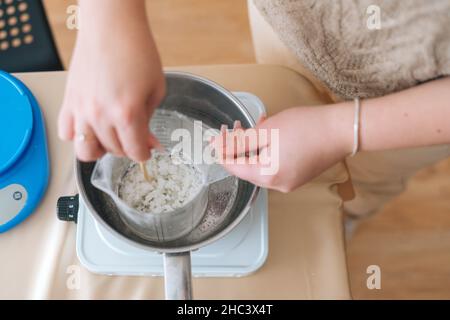 High-angle view of female artisan mixing ingredients for hand candle-making in glass jar into pot of boiling water for creating candle building Stock Photo