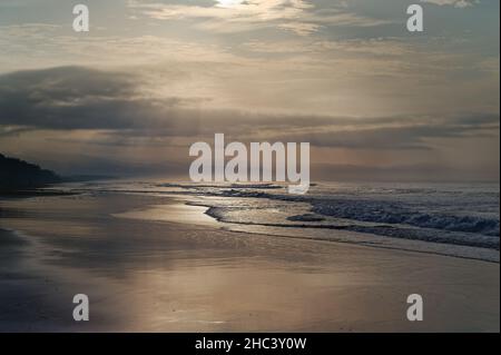 After sunrise , stunning light rays breaking through the dark clouds , give a wonderful metallic reflection on the crashing waves of this Bali beach . Stock Photo