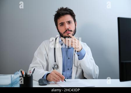 Young doctor looking at camera and thinking sitting at desk in a medical office. Stock Photo