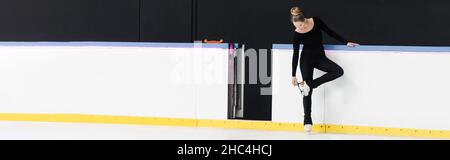 full length of figure skater in black bodysuit checking blade on ice skates near frozen ice arena, banner Stock Photo