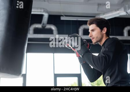 Side view of sportsman in boxing gloves looking at blurred punching bag in gym Stock Photo