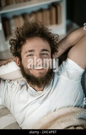 Handsome Guy with Blue Eyes, Curly Hair, a Beard and a Wide Smile Lies Under the Cover in his Bed. Top View. Close-up Portrait. Book Shelves Background. High quality photo Stock Photo