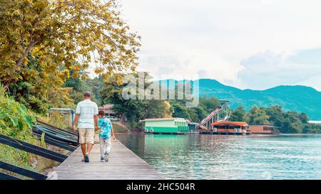 Back view of Asian grandfather and grandchild wear protective face mask to prevent Coronavirus (COVID-19) walking in a nature path on wooden bridge al Stock Photo