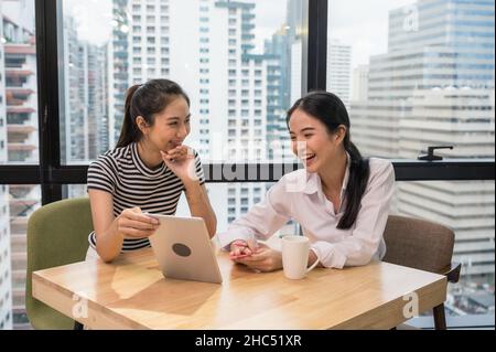 Two asian female employees having fun talking and showing tablet on desk in modern office Stock Photo