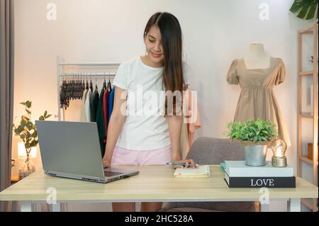 Asian woman entrepreneur of online clothing shop standing using laptop and checking inventory on desk in home office Stock Photo