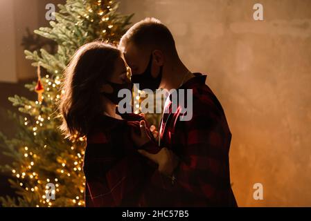 A young couple man and woman in disposable medical masks under Christmas tree. Stock Photo