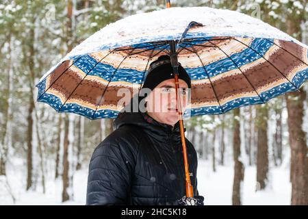 Portrait of young man in warm hat outside on rural winter snowy background holding umbrella. Happy millennial serious outdoor, cold weather. Caucasian Stock Photo