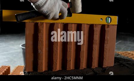 Industrial background, worker in gloves using hammer for laying brick in cement. Close up of a man using level and hammer to make a straight row of br Stock Photo