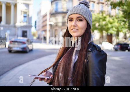 Happy young female millennial with long brown hair in warm knitted hat and stylish leather jacket smiling and looking away while standing on city street, with smartphone in hand Stock Photo