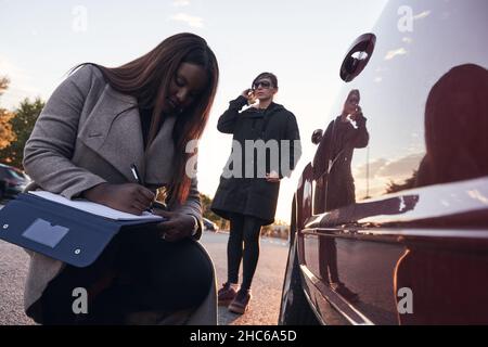 insurance agent examines the car while the driver talks on the mobile phone. Accident claim process. African-American woman checking the damage to the Stock Photo