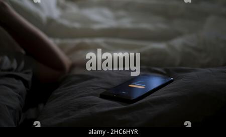 Close up of sleeping man being woken up by his phone lying on the bed. Male hand trying to turn off the alarm clock lying near on a blanket at home ea Stock Photo
