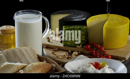 Still life in the old style with a jug and a mug of milk, a heads of cheese, toast, honey and eggs on black background. Cheese camembert, blue cheese, Stock Photo