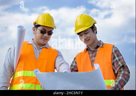 Two Asian engineers or technicians wear a yellow helmet with an orange reflective jacket and stand to hold a blueprint with a smile. Both consult the Stock Photo