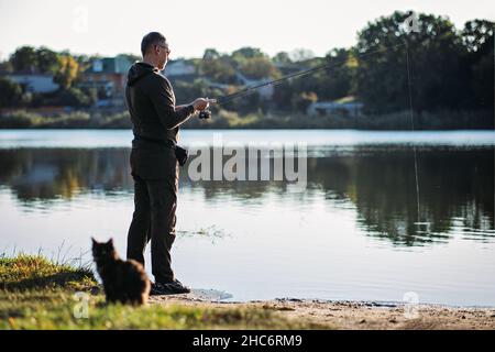 Fisherman with spinning rod and cat near him on nature background. Angler man with fishing spinning or casting rod by the river. Fisherman with rod Stock Photo
