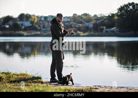 Fisherman with spinning rod and cat near him on nature background. Angler man with fishing spinning or casting rod by the river. Fisherman with rod Stock Photo