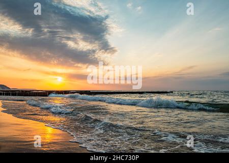 Sunset in the wavy sea with a wooden breakwater by the sandy shore Stock Photo