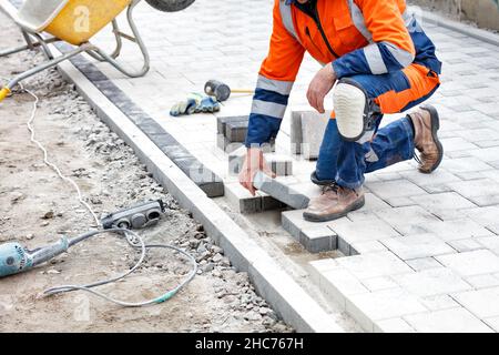 A worker in blue and orange overalls lays paving slabs on a pedestrian walkway under construction. Selective focus, copy space. Stock Photo