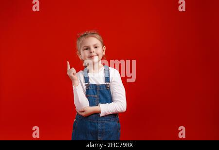 a little schoolgirl in a denim dress poses on a red isolated background. Stock Photo