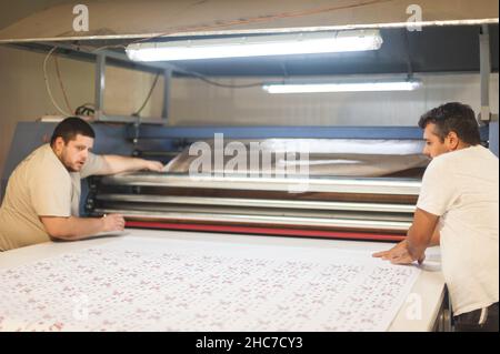 Two technician workers operator works on large premium fabric industrial textile sublimation roller heat press printing machine in digital printshop o Stock Photo