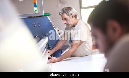 Two technician workers operator works on large premium fabric industrial textile sublimation roller heat press printing machine in digital printshop o Stock Photo