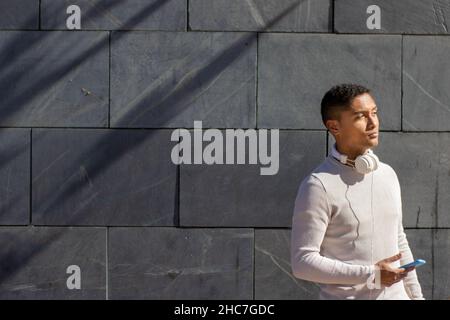 Portrait of a black man with headphones, using your mobile phone. A street gray brick wall in the background. Music, students and black people concept Stock Photo