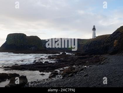The black cobblestone beach and volcanic rock boulders with Yaquina Head lighthouse atop a cliff in the background. Photographed on the Oregon coast. Stock Photo