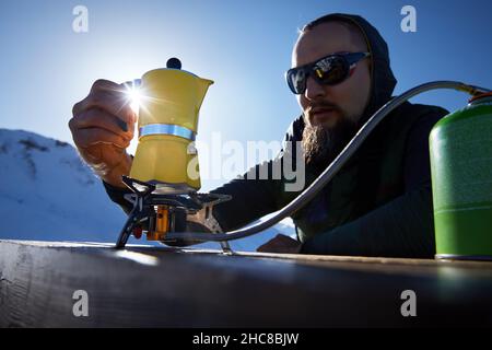 Bearded man hiker making coffee from Yellow Moka mocha pot on gas stove outdoors in the snow winter mountains. Old style coffee vintage pot outdoor ca Stock Photo