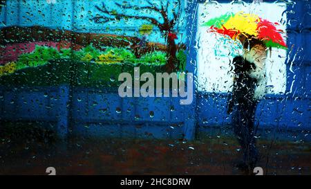 Raindrops on a glass window cause a blurred view of a winter scene of a human figure with a colourful umbrella Stock Photo