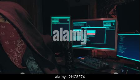 Hacker in a hood sits in front of computer screens and hacks databases Stock Photo