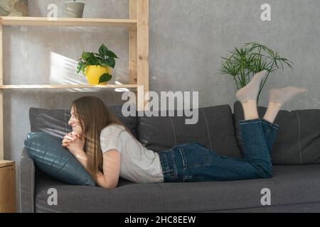 Teenage girl lies on couch and looks dreamily in front of her. Young woman is resting on sofa in living room. Front view. Stock Photo