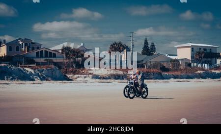 Daytona Beach, Florida, USA - December 17, 2021: Mature caucasian couple ride rental bicycles on beach with luxury homes and pretty blue sky Stock Photo