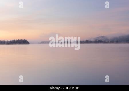 Early winter morning during a vibrant sunrise full of pastel colors and a veil of fog over the lake in Maastricht.  The still water creates reflection Stock Photo