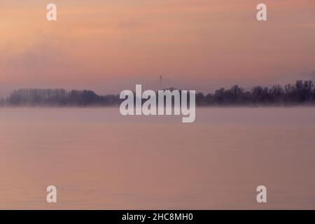 Early winter morning during a vibrant sunrise full of pastel colors and a veil of fog over the lake in Maastricht.  The still water creates reflection Stock Photo