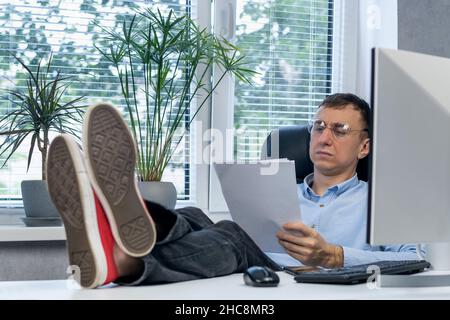 Young director is sitting in his office in leather chair, reads documents and puts feet on table. Man worker. Stock Photo