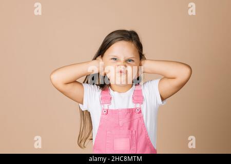 Portrait photo of child closing ears with hands not hear what others say with frowny-face with shining eyes looking at camera wearing bright pink Stock Photo