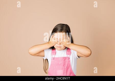 Close up portrait rebellious kid closing eyes with hands not see others with closed mouth not talk with others wearing bright pink jumpsuit and white Stock Photo