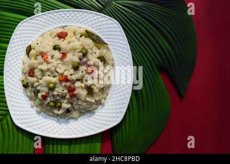 Upma Indian semolina breakfast healthy dish served in white plate. Top view of vegetable Upama on red and leaf background, space to write font text. Stock Photo