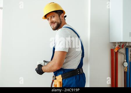 Confident electrician in hard hat and protective gloves smiling at camera while installing electrical socket in a room Stock Photo