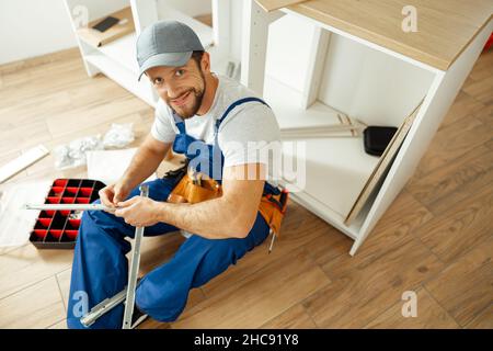 High angle view of handyman in uniform smiling at camera while sitting on the floor and assembling drawer in kitchen Stock Photo