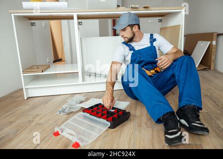 Focused handyman in overalls sitting on the floor while assembling kitchen cabinet in apartment Stock Photo