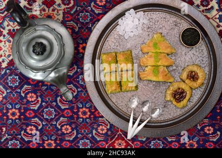 Fresh baklava, oriental pastry made from puff pastry and nuts, lies on a copper tray that stands next to an old teapot on an old oriental carpet, phot Stock Photo