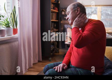 Frustrated senior man in red sweater holding hand on his neck sitting on sofa Stock Photo