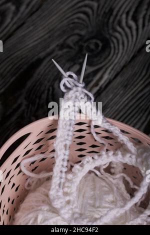 Knitting with needles. A skein of white thread and a fragment of a knitted product. Lie in a basket. On black pine boards. Close-up. Stock Photo