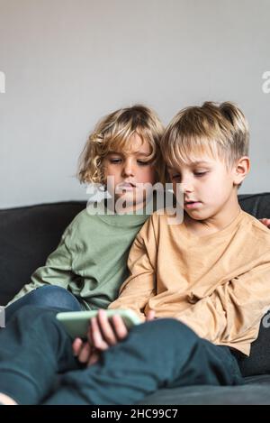 Serious little sister and brother in casual apparel with blond hair sharing mobile phone while spending weekend together sitting on couch at home Stock Photo