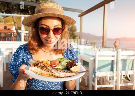 Happy woman with baked sea bass fish in a seafood restaurant. The Mediterranean diet is an indispensable source of omega-3 fats and vitamins Stock Photo