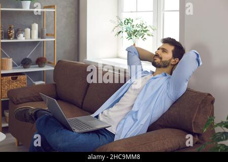 Happy young man thinking and dreaming while relaxing on comfortable couch with laptop Stock Photo