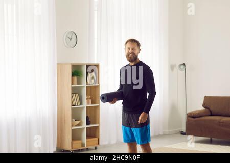 Happy fit man with wireless earbuds standing in living room, holding yoga mat and smiling Stock Photo