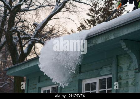 Ice dam prevention. Man holding shovel cleaning roof from snow, ice and icicles during wintertime Stock Photo