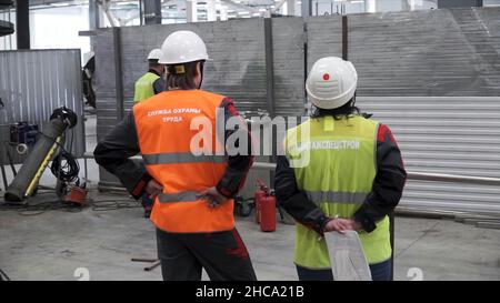 Rear view of workers talking while walking in shipping yard. Back view of two men in white safety hard hats are standing together in construction area Stock Photo