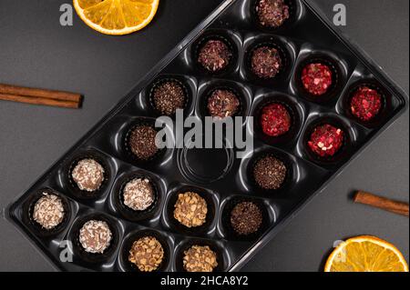 Colorful chocolate truffles in a candy box with cinnamon and dried oranges on a black background. Top view. Stock Photo